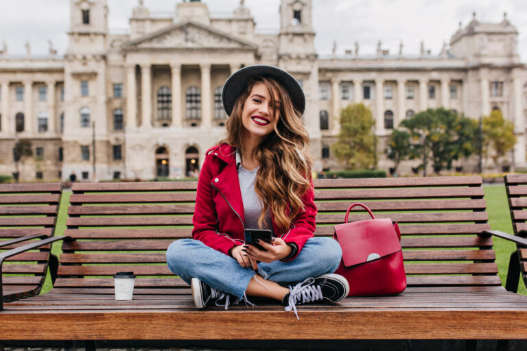 Spectacular lady with trendy hairstyle sitting on steps in front of old historical building. Romantic female traveler with red backpack posing with smartphone, enjoying sightseeing.